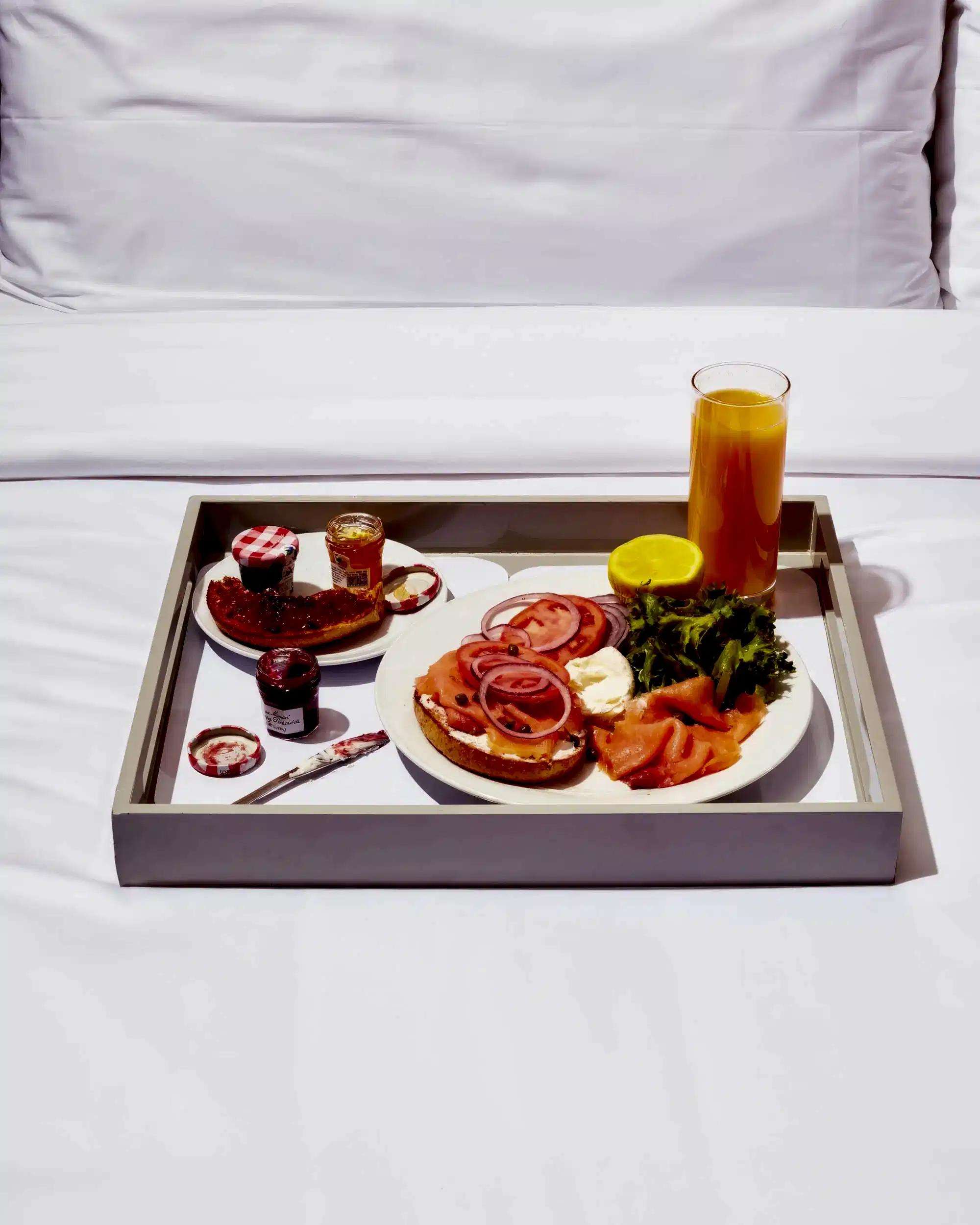 An in-room dining breakfast tray of a smoked salmon bagel, jams, and orange juice on a bed.